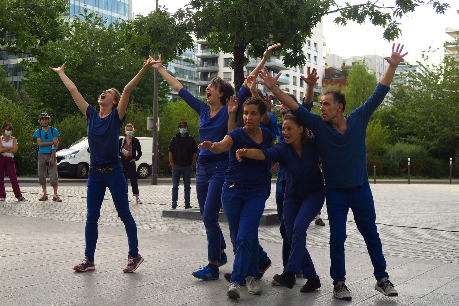 5 danseurs font des gestes avec leurs bras vers le haut. Ils sont habillés en bleu, et sont dans l'espace public